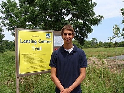 Dave Miller at the entrance to the Lansing Center Trail. The tree line in the distance is the railroad bed he will be clearing and preparing as an addition to the trail. davemiller_bytrail400
